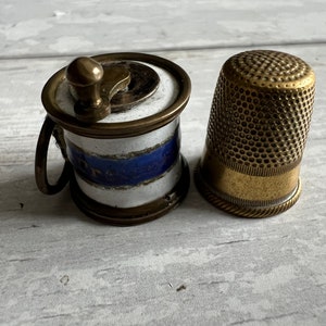 May include: A vintage sewing kit featuring a small, brass thimble and a miniature thread winder. The winder has a white and blue striped ceramic body with brass accents. The thimble is gold-toned with a textured surface.