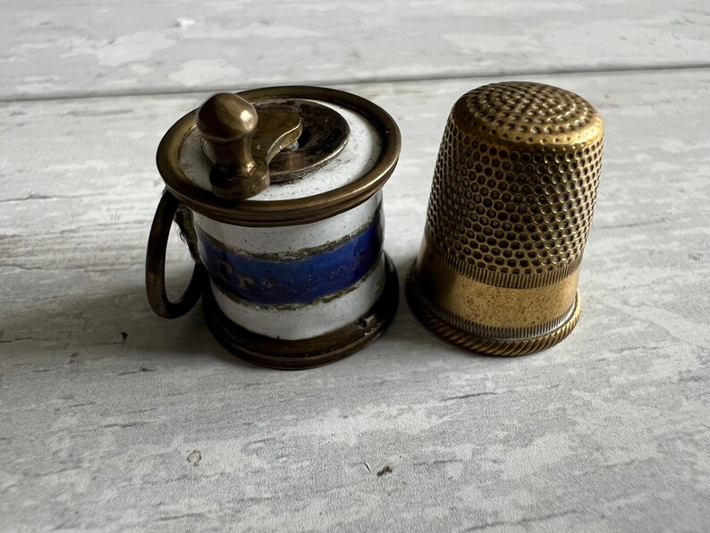 May include: A vintage sewing kit featuring a small, brass thimble and a miniature thread winder. The winder has a white and blue striped ceramic body with brass accents. The thimble is gold-toned with a textured surface.