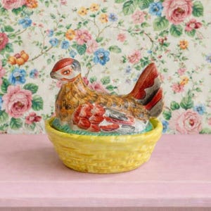 May include: A ceramic hen figurine with a red comb and tail, in a yellow basket. The hen has brown, red, and white details. The background features floral wallpaper with pink, blue, and orange flowers.