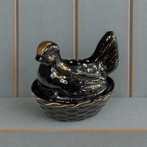 May include: A black ceramic hen on a nest figurine featuring gold painted accents on the head, wings, and basket base. The bird has white dot details on its back and a glossy finish. This decorative lidded container is shaped like a sitting hen.