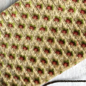 Pode incluir: Um close-up de um tecido cor de creme com um padr&atilde;o repetitivo de pequenas flores vermelhas e verdes. O tecido tem uma superf&iacute;cie texturizada, quase felpuda.