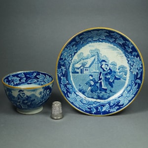May include: A blue and white porcelain teacup and saucer set with a floral border and a scene of people walking in a landscape. The saucer has a gold rim.