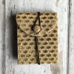 Pode incluir: Uma pequena bolsa de tecido bege com fecho de cord&atilde;o castanho e um &uacute;nico bot&atilde;o. A bolsa tem um padr&atilde;o floral com pequenas flores vermelhas.