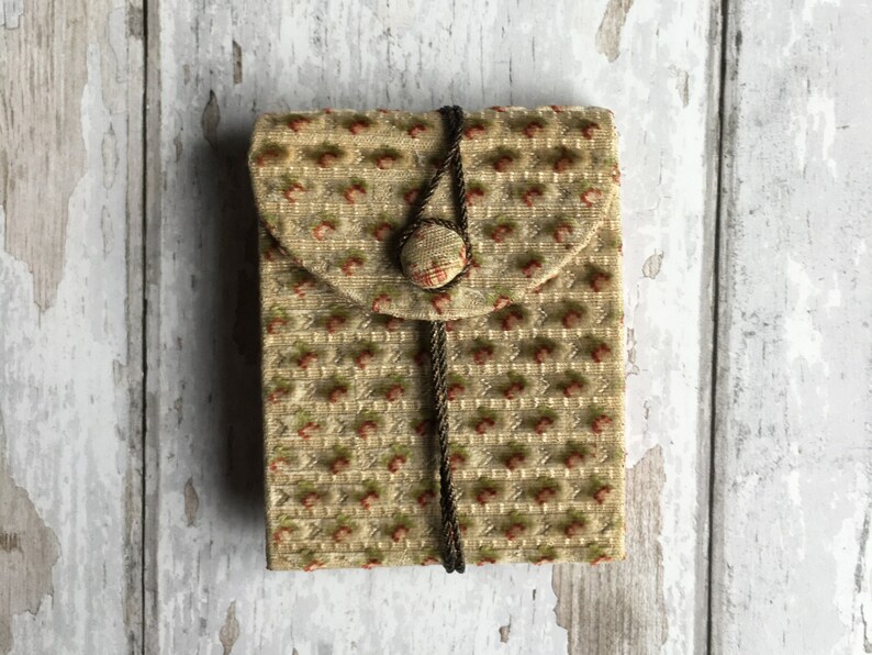 Pode incluir: Uma pequena bolsa de tecido bege com fecho de cord&atilde;o castanho e um &uacute;nico bot&atilde;o. A bolsa tem um padr&atilde;o floral com pequenas flores vermelhas.