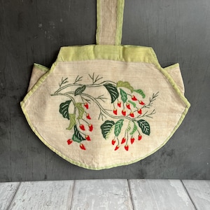 May include: A vintage, off-white linen bag with a green embroidered floral design. The bag has a green fabric handle and a scalloped edge.