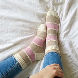 May include: A pair of striped socks in pink and beige with a white background. The socks are worn on a person's feet, with the person's legs crossed and wearing blue jeans.