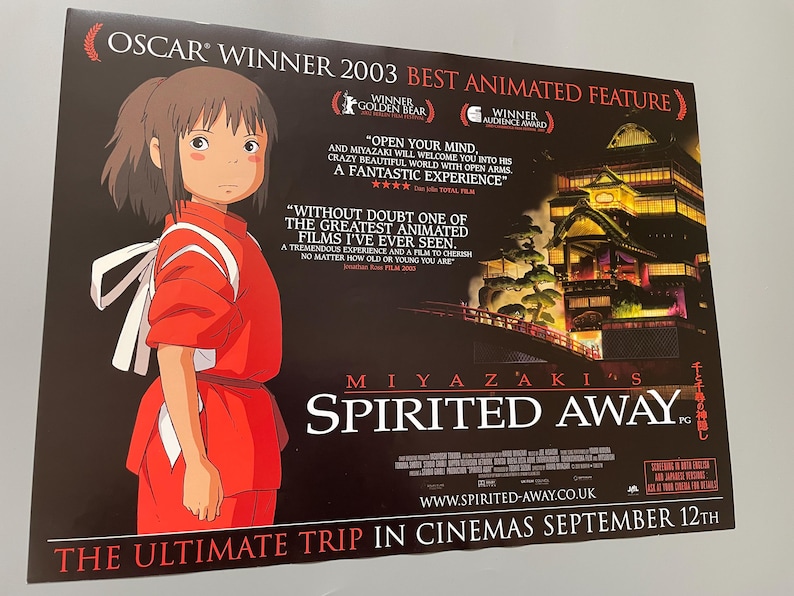May include: A movie poster for Miyazaki's "Spirited Away," featuring a young girl in a red outfit. The poster includes text about the film's awards and release date, with a dark background and a Japanese-style building.