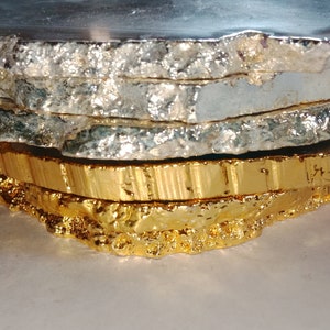 May include: Set of four agate coasters with a natural, irregular shape. The top coaster is a dark grey color, the next two are silver, and the bottom coaster is gold.