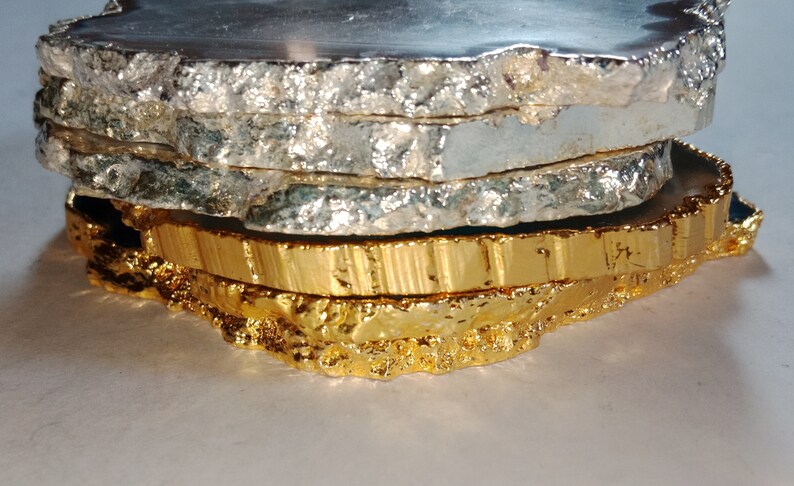May include: Set of four agate coasters with a natural, irregular shape. The top coaster is a dark grey color, the next two are silver, and the bottom coaster is gold.