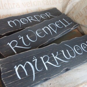Pu&ograve; includere: Tre cartelli di legno dipinti di nero con lettere bianche. I cartelli recitano "Mordor", "Rivendell" e "Mirkwood".