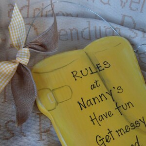 K&ouml;nnte beinhalten: Gelbes Holzschild in Form von Gummistiefeln mit dem Text "RULES at Nanny's Have fun Get messy" darauf bemalt. Eine braune und wei&szlig;e Karobinde ist oben am Schild befestigt.