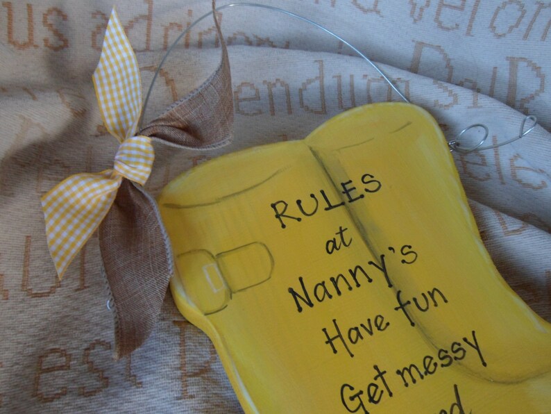 K&ouml;nnte beinhalten: Gelbes Holzschild in Form von Gummistiefeln mit dem Text "RULES at Nanny's Have fun Get messy" darauf bemalt. Eine braune und wei&szlig;e Karobinde ist oben am Schild befestigt.