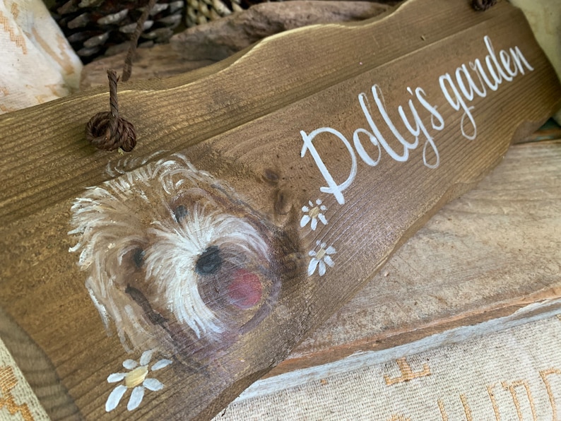 K&ouml;nnte beinhalten: Ein Holzschild mit einem gemalten Hund und dem Text "Polly's garden". Das Schild ist braun mit wei&szlig;en Farbakzenten und hat ein Seil zum Aufh&auml;ngen.