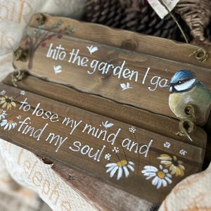 K&ouml;nnte beinhalten: Zwei Holzschilder mit wei&szlig;er Aufschrift und Blumendesigns. Das obere Schild sagt "Into the garden I go" und zeigt einen blauen Vogel. Das untere Schild sagt "To lose my mind and find my soul".