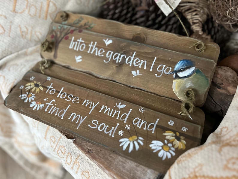 K&ouml;nnte beinhalten: Zwei Holzschilder mit wei&szlig;er Aufschrift und Blumendesigns. Das obere Schild sagt "Into the garden I go" und zeigt einen blauen Vogel. Das untere Schild sagt "To lose my mind and find my soul".
