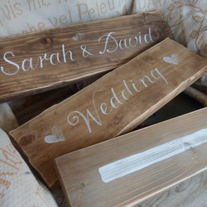 Pu&ograve; includere: Cartelli di nozze in legno con scritte bianche. Un cartello recita "Sarah & David", un altro dice "Wedding", e un terzo ha una freccia bianca. I cartelli sono in varie tonalit&agrave; di marrone e beige.