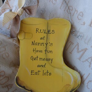 K&ouml;nnte beinhalten: Gelbes Holzschild in Form eines Gummistiefels mit dem Text "RULES at Nanny's Have fun Get messy and Eat lots" in Schwarz.