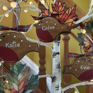 May include: Three wooden robin ornaments with red chests and white painted details, each personalized with the names Katie, Chloe, and Evie. Each bird features a green and red plaid ribbon bow and hangs alongside stained glass leaf ornaments.