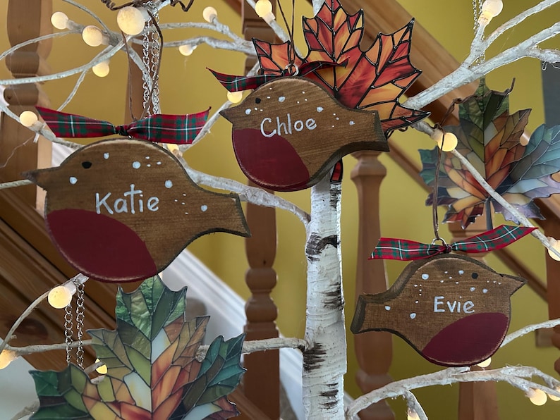May include: Three wooden robin ornaments with red chests and white painted details, each personalized with the names Katie, Chloe, and Evie. Each bird features a green and red plaid ribbon bow and hangs alongside stained glass leaf ornaments.