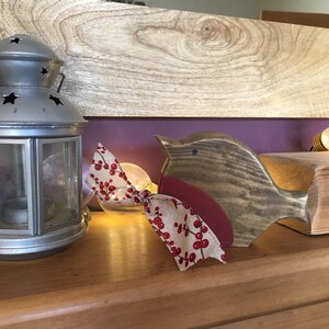 May include: A silver-colored metal lantern with star-shaped cutouts sits next to a wooden bird figurine. The bird has a dark wood body, a red chest, and a beige ribbon tied around its neck featuring a red berry and leaf pattern.