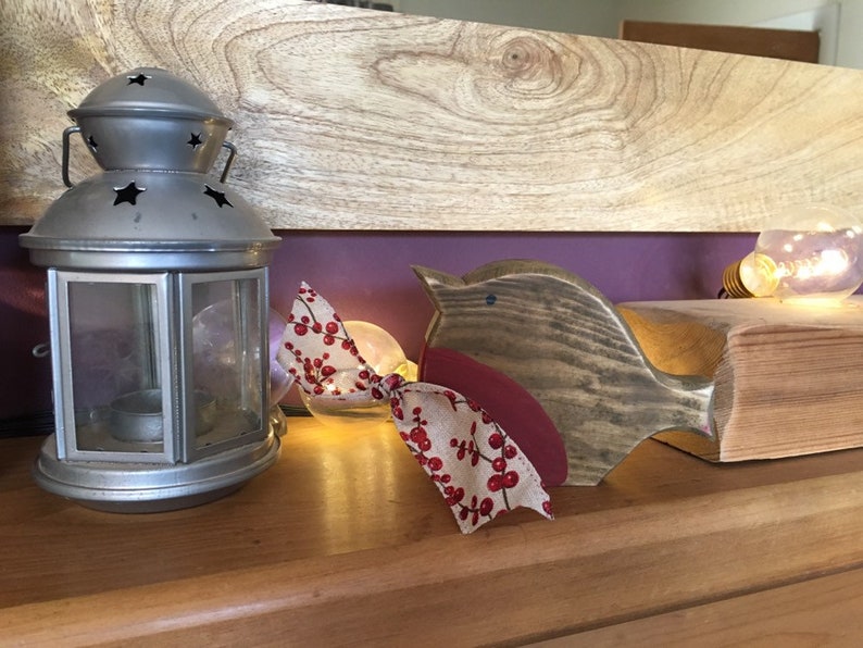 May include: A silver-colored metal lantern with star-shaped cutouts sits next to a wooden bird figurine. The bird has a dark wood body, a red chest, and a beige ribbon tied around its neck featuring a red berry and leaf pattern.