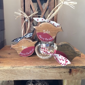 May include: Three wooden bird ornaments with red painted chests, each featuring the text Merry Christmas and a name, Ben or Isla. The birds are decorated with raffia, plaid, and berry-patterned ribbons, and one sits atop a small glass jar.