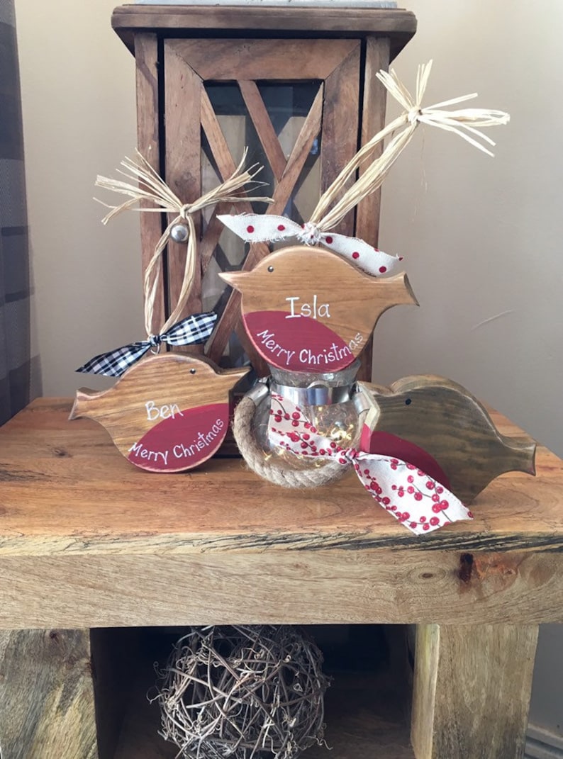 May include: Three wooden bird ornaments with red painted chests, each featuring the text Merry Christmas and a name, Ben or Isla. The birds are decorated with raffia, plaid, and berry-patterned ribbons, and one sits atop a small glass jar.