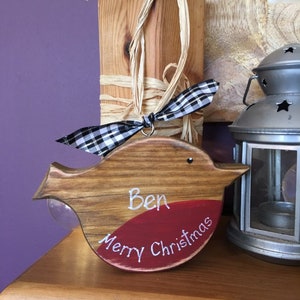 May include: A wooden bird-shaped ornament with a natural wood grain finish and a painted red chest. The ornament features the white hand-painted text "Ben" and "Merry Christmas" and is decorated with a black and white checkered ribbon.