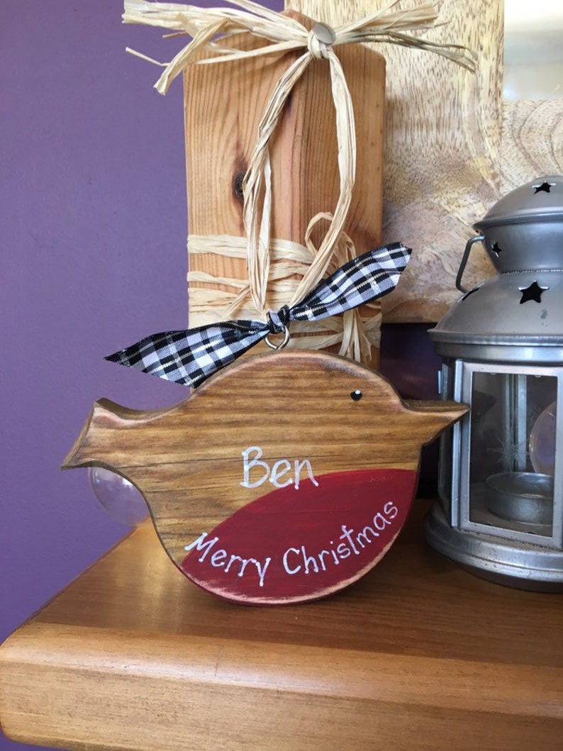 May include: A wooden bird-shaped ornament with a natural wood grain finish and a painted red chest. The ornament features the white hand-painted text "Ben" and "Merry Christmas" and is decorated with a black and white checkered ribbon.