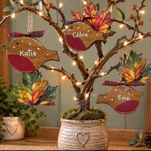 May include: Three wooden bird ornaments with red chests, each personalized with the names Katie, Chloe, and Evie, hanging from a lit tree alongside two colorful autumn leaf ornaments, all accented with small red and green plaid ribbons.