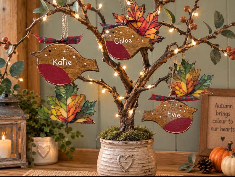 May include: Three wooden bird ornaments with red chests, each personalized with the names Katie, Chloe, and Evie, hanging from a lit tree alongside two colorful autumn leaf ornaments, all accented with small red and green plaid ribbons.