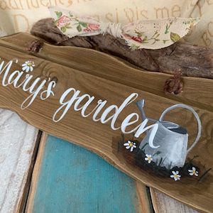 K&ouml;nnte beinhalten: Ein Holzschild mit den Worten "Mary's garden" in wei&szlig;er Farbe. Das Schild ist mit einem bemalten Gie&szlig;kannen und G&auml;nsebl&uuml;mchen verziert.