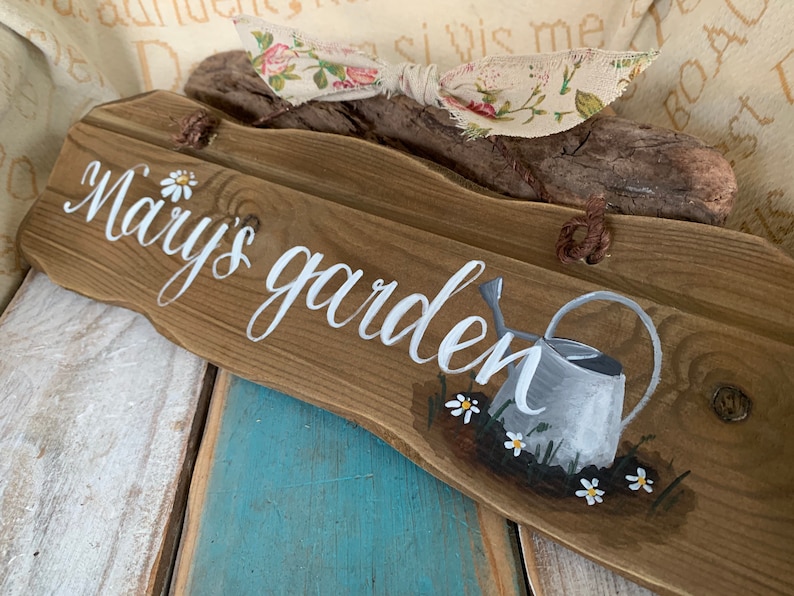 K&ouml;nnte beinhalten: Ein Holzschild mit den Worten "Mary's garden" in wei&szlig;er Farbe. Das Schild ist mit einem bemalten Gie&szlig;kannen und G&auml;nsebl&uuml;mchen verziert.
