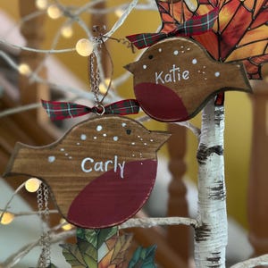 May include: Two wooden robin ornaments with brown bodies and red chests, each featuring white painted dots and the names Carly and Katie written in white script. Each bird is topped with a small green and red plaid ribbon bow.