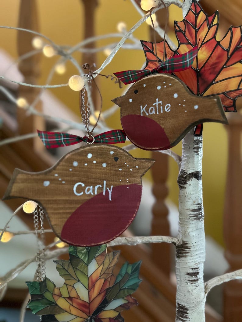 May include: Two wooden robin ornaments with brown bodies and red chests, each featuring white painted dots and the names Carly and Katie written in white script. Each bird is topped with a small green and red plaid ribbon bow.