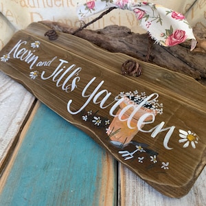 K&ouml;nnte beinhalten: Ein rustikales Holzschild mit dem Text "Kevin and Jill's Garden" in wei&szlig;er Farbe. Das Schild ist mit wei&szlig;en G&auml;nsebl&uuml;mchen und einem gemalten Bild eines Blumentopfs mit Blumen und Gartenger&auml;ten verziert.