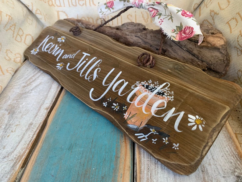 K&ouml;nnte beinhalten: Ein rustikales Holzschild mit dem Text "Kevin and Jill's Garden" in wei&szlig;er Farbe. Das Schild ist mit wei&szlig;en G&auml;nsebl&uuml;mchen und einem gemalten Bild eines Blumentopfs mit Blumen und Gartenger&auml;ten verziert.