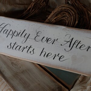 Pu&ograve; includere: Un cartello rettangolare in legno bianco con la frase "Happily Ever After starts here" in corsivo nero. Il cartello ha un aspetto invecchiato con venature del legno visibili e un cuore. Arredamento rustico.