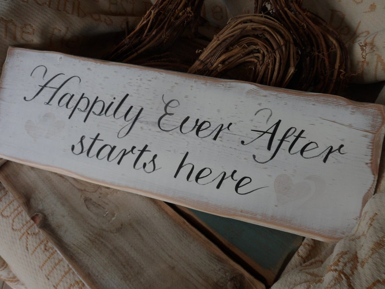 Pu&ograve; includere: Un cartello rettangolare in legno bianco con la frase "Happily Ever After starts here" in corsivo nero. Il cartello ha un aspetto invecchiato con venature del legno visibili e un cuore. Arredamento rustico.