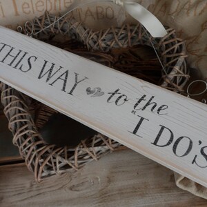 Pu&ograve; includere: Un cartello in legno bianco sporco con la scritta "THIS WAY to the I DO'S" in nero. Il cartello a forma di freccia presenta un piccolo disegno a forma di cuore. Sullo sfondo, una decorazione a forma di cuore intrecciata.