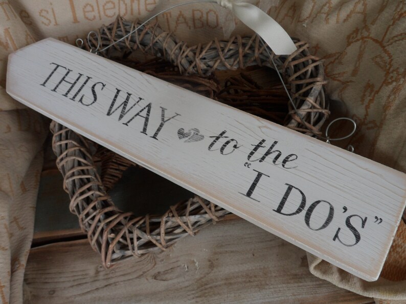 Pu&ograve; includere: Un cartello in legno bianco sporco con la scritta "THIS WAY to the I DO'S" in nero. Il cartello a forma di freccia presenta un piccolo disegno a forma di cuore. Sullo sfondo, una decorazione a forma di cuore intrecciata.