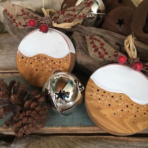 Pode incluir: Dois ornamentos redondos de madeira pintados para parecer um pudim de Natal com cobertura branca e granulado marrom. Os ornamentos estão pendurados em um barbante com bagas vermelhas. Um sino de prata está em primeiro plano.