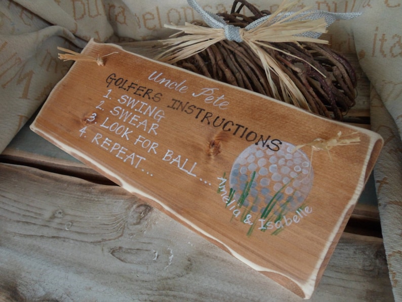 Puede incluir: Un letrero de madera con el texto "Uncle Pete Golfers Instructions" y una lista de instrucciones: "1. Swing", "2. Swear", "3. Look for Ball", "4. Repeat". El letrero tambi&eacute;n presenta una bola de golf pintada y el texto "Amelia & Isabelle".