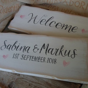 Pu&ograve; includere: Due cartelli rustici in legno bianco. Uno dice "Welcome" in corsivo nero con un cuore rosa. L'altro dice "Sabina & Markus 1st September 2018" con un cuore rosa. I cartelli hanno un aspetto invecchiato.