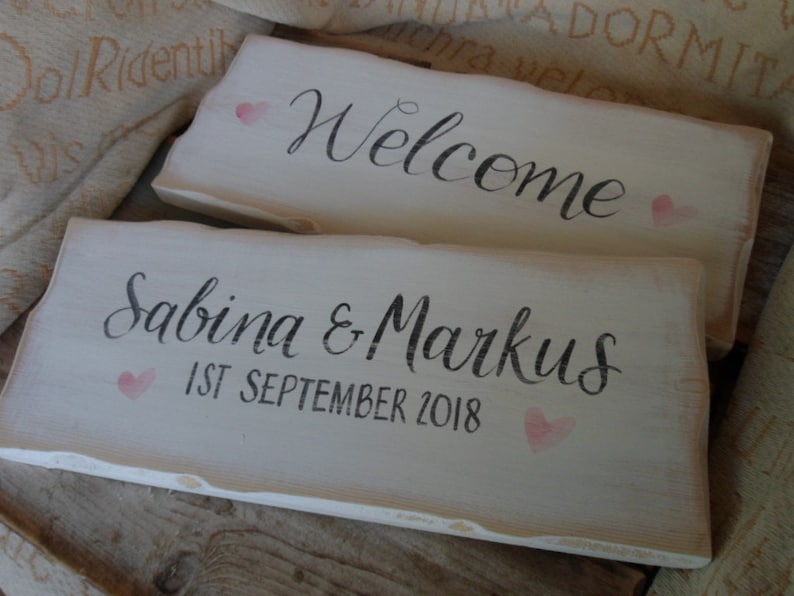 Pu&ograve; includere: Due cartelli rustici in legno bianco. Uno dice "Welcome" in corsivo nero con un cuore rosa. L'altro dice "Sabina & Markus 1st September 2018" con un cuore rosa. I cartelli hanno un aspetto invecchiato.