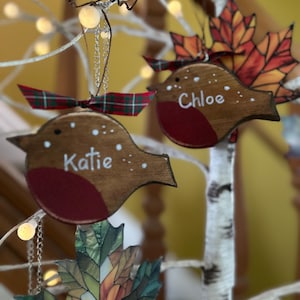 May include: Two wooden robin ornaments with red breasts and brown bodies, each featuring a white name, Katie and Chloe, painted on the side. Each bird is topped with a small green and red plaid ribbon and hangs from a silver chain.
