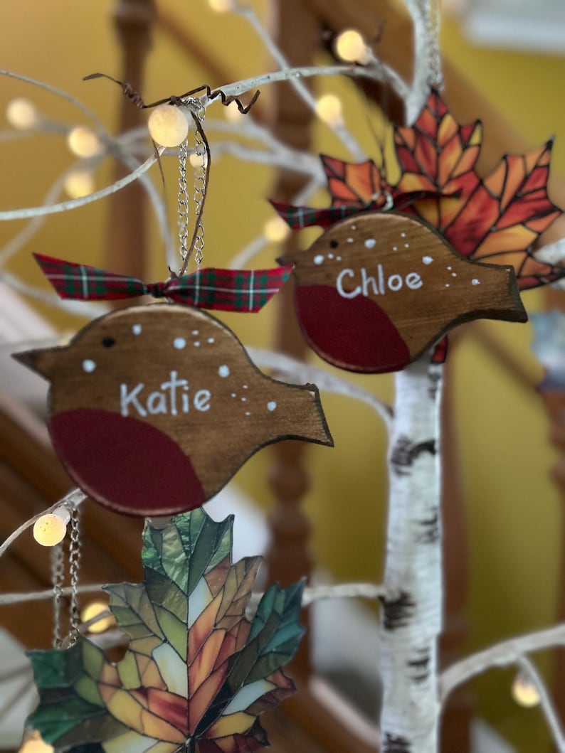May include: Two wooden robin ornaments with red breasts and brown bodies, each featuring a white name, Katie and Chloe, painted on the side. Each bird is topped with a small green and red plaid ribbon and hangs from a silver chain.