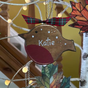 May include: A wooden bird-shaped ornament with a dark brown finish, a red chest, and white painted dots. The name Katie is written in white script across the center. A red and green plaid ribbon is tied to the top silver hanging chain.
