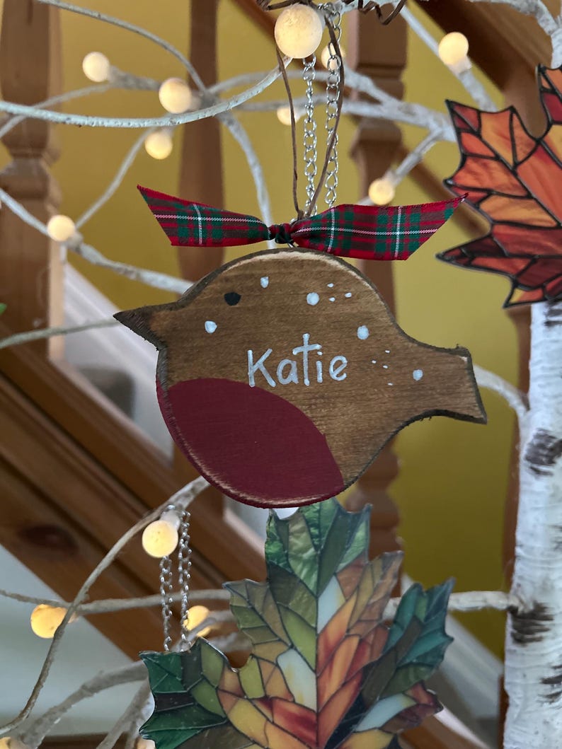 May include: A wooden bird-shaped ornament with a dark brown finish, a red chest, and white painted dots. The name Katie is written in white script across the center. A red and green plaid ribbon is tied to the top silver hanging chain.