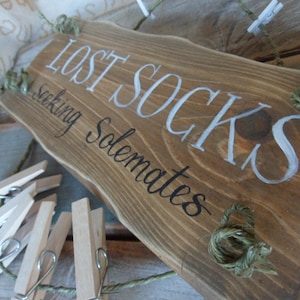 Pode incluir: Uma placa de madeira rústica com o texto "Lost Socks Seeking Solemates" em letras brancas. A placa tem um aspecto envelhecido e é decorada com barbante. Há prendedores de roupa de madeira pendurados no barbante.